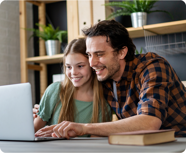 Smiling father and daughter sitting together at a desk, looking at a laptop screen in a cozy room