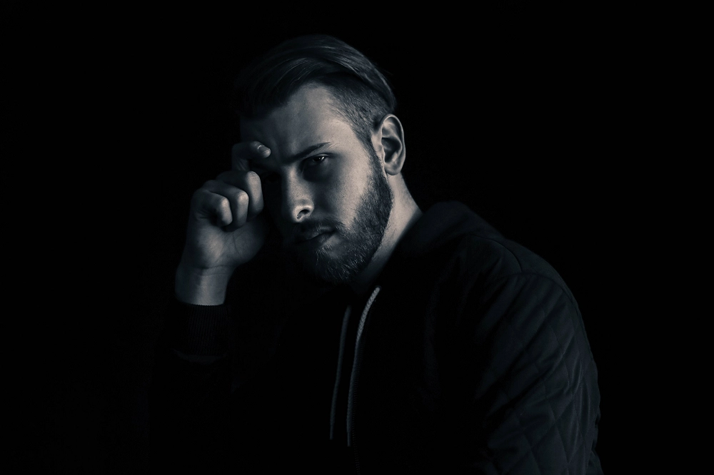 Man in dark lighting with a serious expression, resting his hand on his forehead in a thoughtful or reflective pose