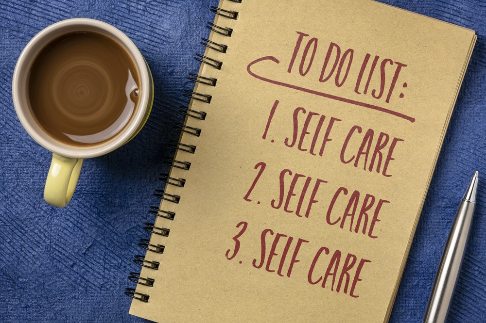 Notebook with “To Do List: 1. Self Care 2. Self Care 3. Self Care” written beside a cup of coffee