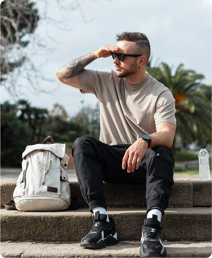 Man sitting outdoors on stone steps with a backpack beside him, shading his eyes from the sun while looking into the distance