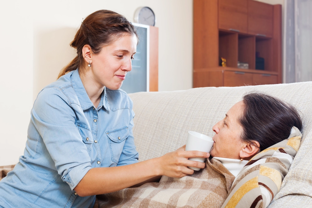 Young woman helping an elderly woman on the couch by offering her a cup to drink from