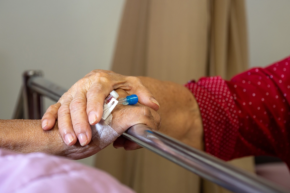 Close-up of a person gently holding the hand of an elderly patient with an IV in a hospital bed